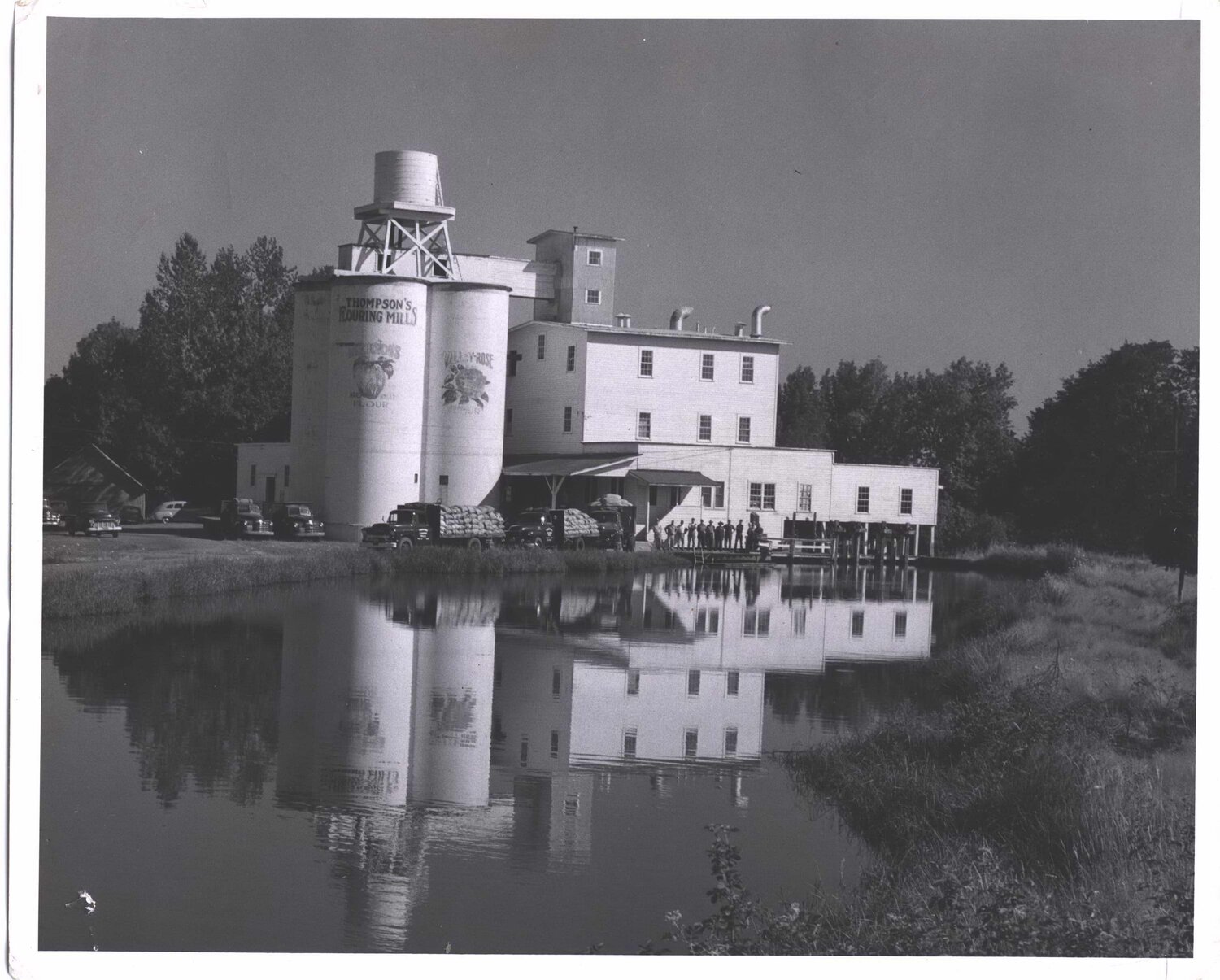 In 1939, a line of trucks filled with flour sacks wait in front of the mill.  People are lined up at the mill's entrance.  The mill is beautifully reflected in the mill pond.